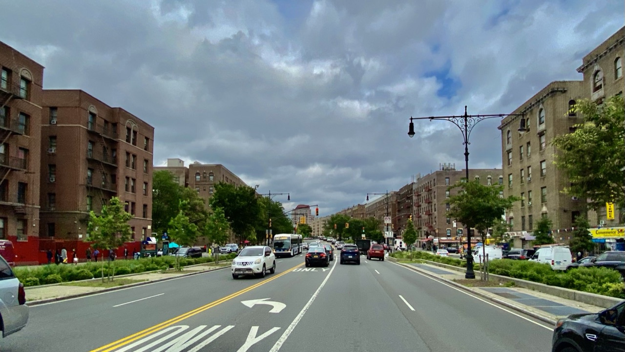 Tree-lined Grand Concourse boulevard in the Bronx with apartment buildings, traffic and ornate street lamps under clouds