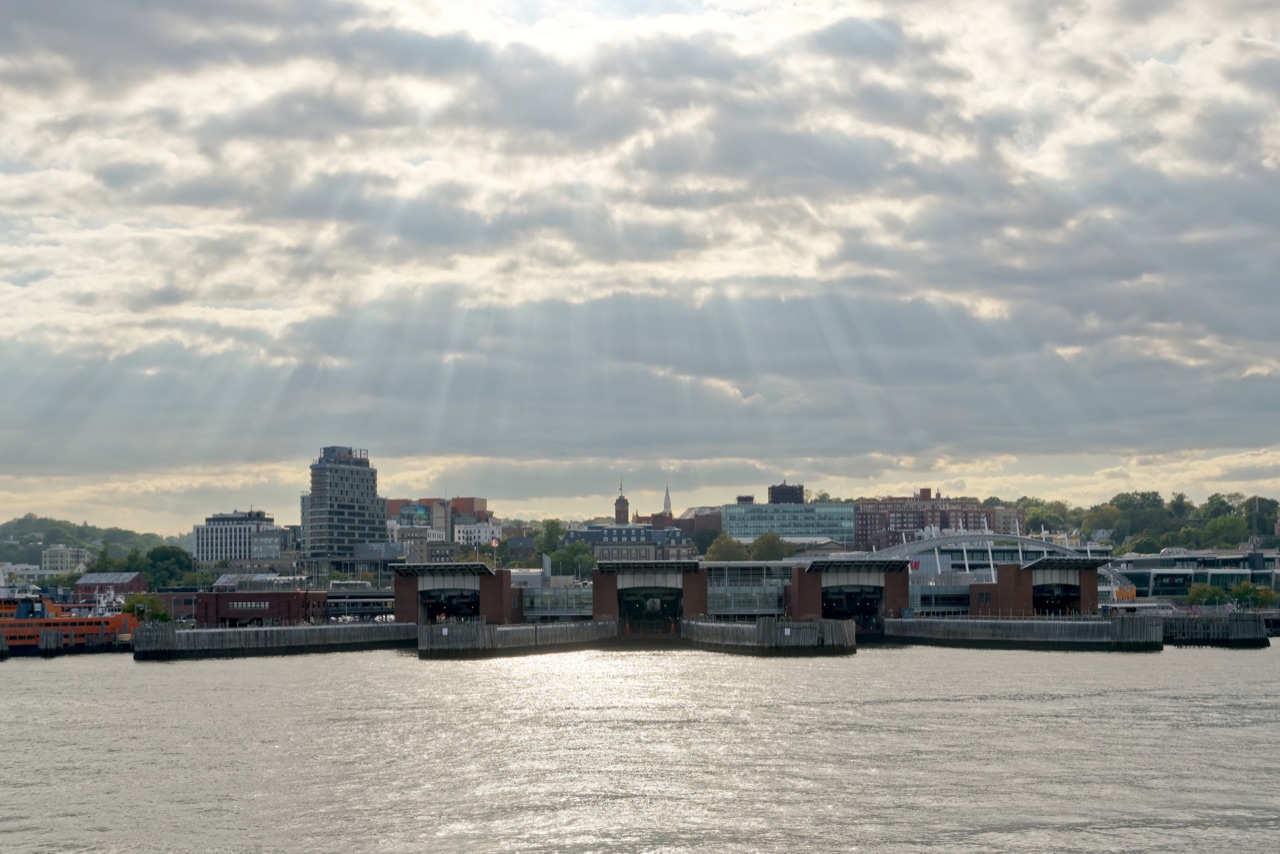 St. George Ferry Terminal and the Staten Island waterfront skyline seen from the water beneath sunbeams through clouds