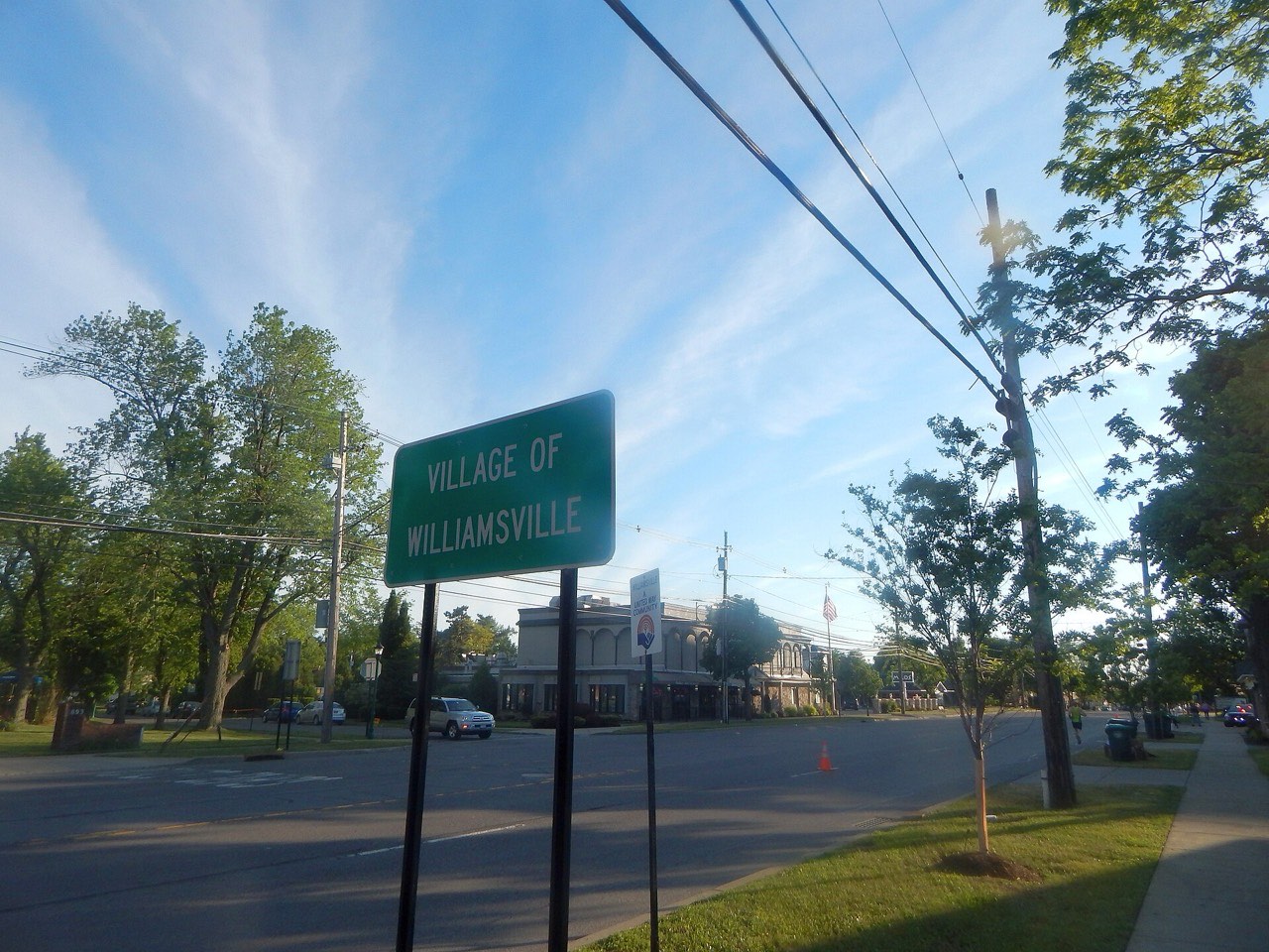 Green Village of Williamsville road sign beside Main Street under a blue sky with trees and buildings